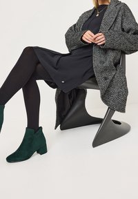 Žena sedící na moderní černé židli, oblečená v černých šatech, šedém herringbone kabátu, černých punčochách a tmavě zelených semišových kotníkových botách.