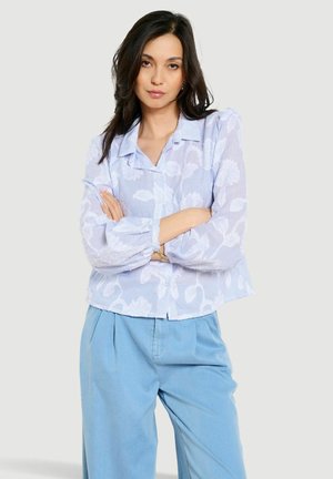 Kobieta o ciemnych włosach, ubrana w jasnoniebieską przejrzystą kwiatową bluzkę i luźne niebieskie spodnie, stojąca z założonymi ramionami na jasnym tle.