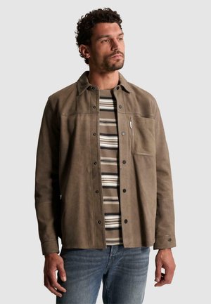 Homme aux cheveux courts et bouclés portant une veste marron à boutons sur une chemise rayée et un jean bleu, debout devant un fond uni.
