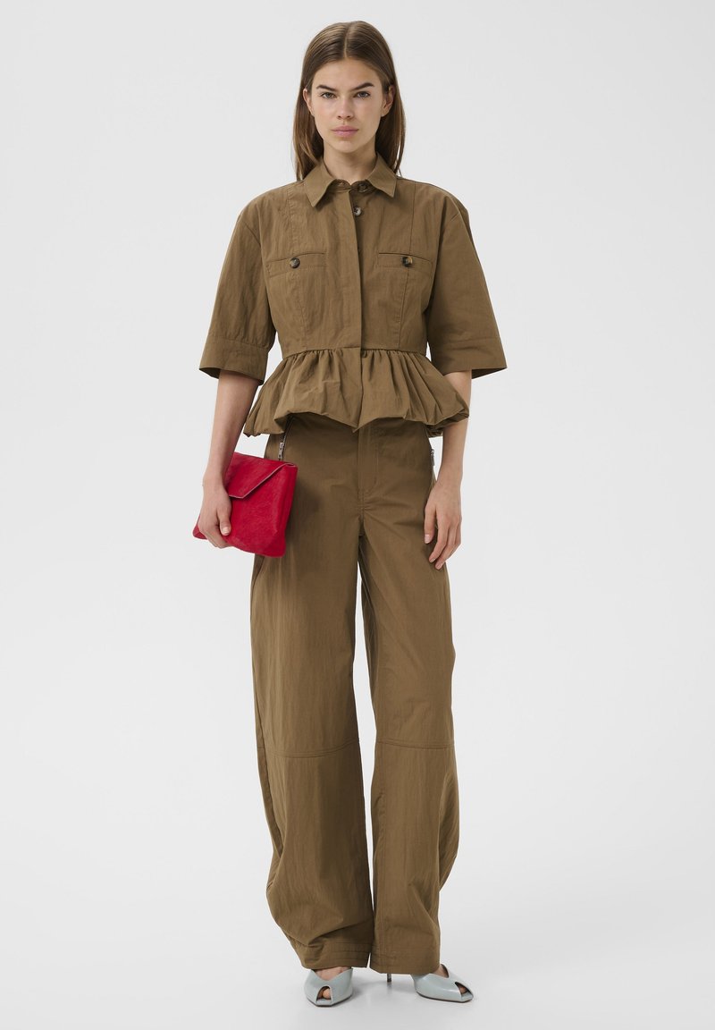 Vrouw draagt een bruin peplumtop met knopen en wijde broek, houdt een rode clutch vast, staat voor een effen lichtgrijze achtergrond.