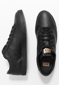 Black leather sneakers with a textured finish, featuring a rubber sole, black laces, and a metallic logo on the tongue. Classic design.