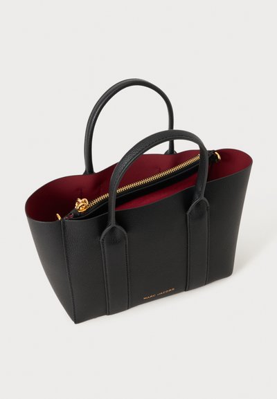 Черна дамска чанта Marc Jacobs с две дръжки, златист цип и вътрешна облицовка в тъмночервен цвят, показана под диагонален ъгъл.