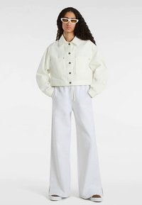 Weiße, kurz geschnittene Jeansjacke mit zwei aufgesetzten Taschen vorne und silbernen Knöpfen, kombiniert mit locker geschnittenen weißen Hosen und weißen Slip-on-Schuhen.