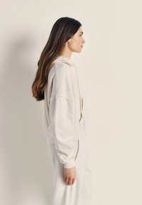 Felpa con cappuccio in pile bianco con tasca a marsupio, polsini a coste e finitura texture, abbinata a pantaloni bianchi coordinati. Modello mostrato di profilo.