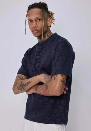 Homme avec des dreadlocks et plusieurs tatouages sur les bras, debout les bras croisés, portant une chemise sombre à manches courtes à motifs et un pantalon de couleur claire.