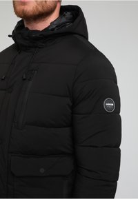 Czarna pikowana kurtka puchowa z zamykaną kieszenią na piersi, zatrzaskami, regulowanym kapturem oraz naszywką z logiem na rękawie. Gładka, pikowana struktura.