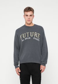 Giovane uomo in maglione grigio con la scritta "FUTURE est. 2018 PARIS", abbinato a pantaloni neri, in piedi su uno sfondo bianco uniforme.