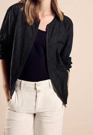 Frau trägt eine schwarze, mit Lochmuster versehene Reißverschlussjacke über einem schwarzen Top, kombiniert mit hoch taillierten, cremefarbenen Hosen mit Knöpfen vorne.