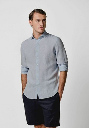 Junger Mann mit hochgekrempelten Ärmeln, der ein hellblaues Hemd mit Knöpfen und dunkle Shorts trägt, steht mit den Händen in den Taschen und blickt zur Seite.