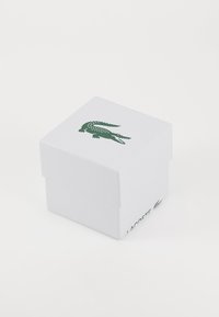 Boîte en papier blanche avec une surface texturée, présentant un logo de crocodile vert sur le dessus et le nom de la marque "Lacoste" sur un côté.
