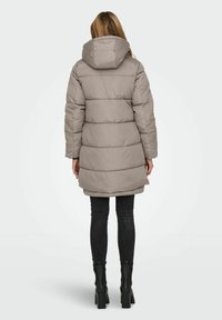 Femme portant un long manteau d'hiver taupe matelassé avec capuche, un jean skinny noir et des bottines à talons noires, vue de dos.