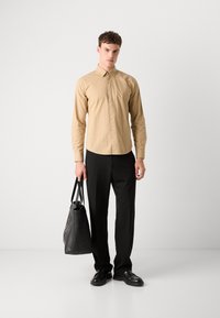Light brown button-up shirt with a collar, paired with black trousers. The model carries a textured black tote bag, wearing black shoes.