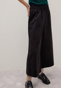 Pantaloni neri a gamba larga con una vita elastica, dotati di tasche laterali e una texture morbida; abbinati a scarpe nere e calzini chiari.