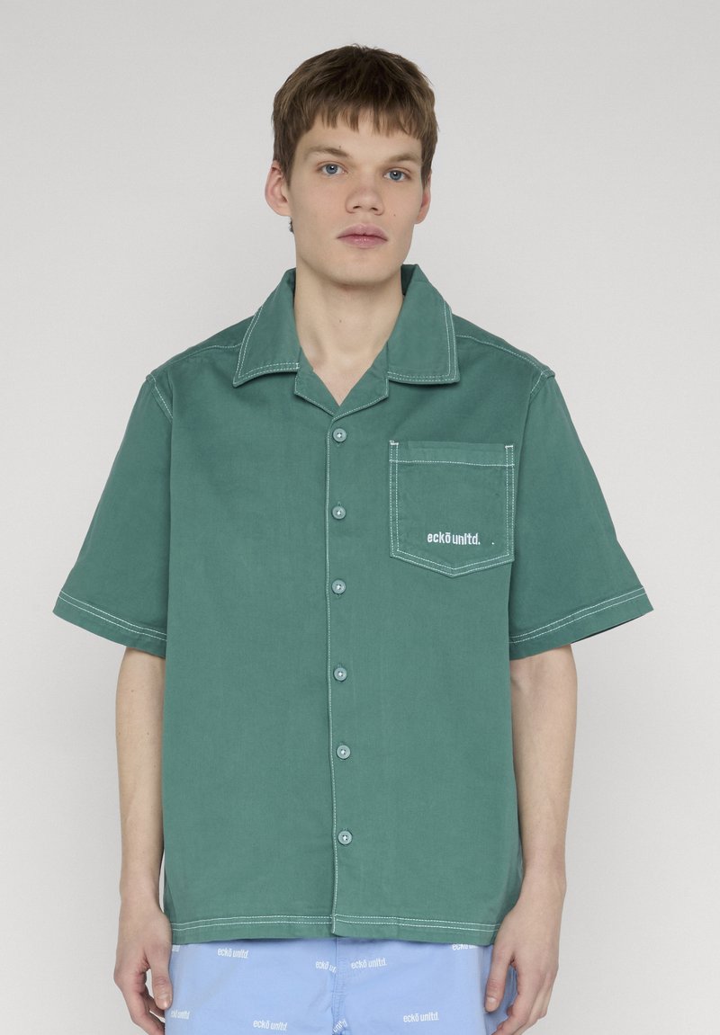 Jeune homme portant une chemise à manches courtes verte avec une poche poitrine et un short bleu à motif de texte blanc.