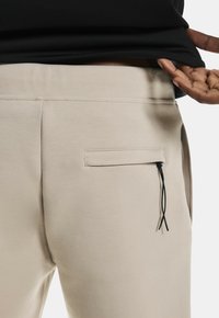 Beige Sweatpants mit glatter Textur, ausgestattet mit einer seitlichen Tasche mit Klappe und schwarzen Kordeln in der Nähe des Bundes.
