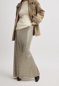 Długa spódnica maxi w złotych cekinach, połączona z kremowym swetrem i beżowym płaszczem, charakteryzująca się rozszerzanym krojem i elegancką fakturą.