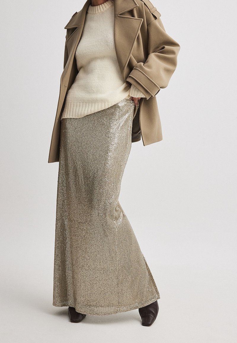 Długa spódnica maxi w złotych cekinach, połączona z kremowym swetrem i beżowym płaszczem, charakteryzująca się rozszerzanym krojem i elegancką fakturą.
