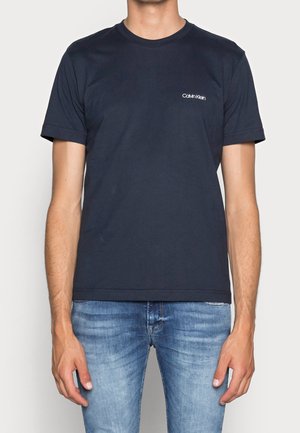 Homme portant un T-shirt uni bleu foncé Calvin Klein et un jean bleu clair, les mains détendues le long du corps.