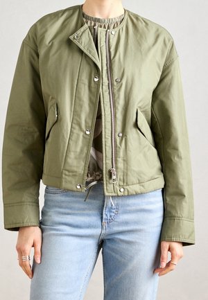 Person iført en olivengrøn jakke med lynlås og trykknapper over en mønstret top med lyseblå jeans, vist fra nakke til hofter.