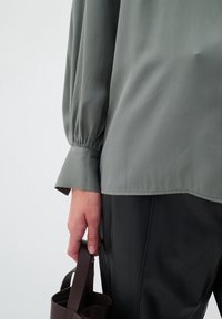 Maglietta grigia a maniche lunghe con texture rigata, con polsini arricciati. Una mano tiene una borsa in pelle marrone. Sotto si vedono dei pantaloni neri.
