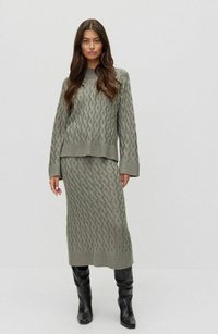 Gehaakt tweedelig set in gedempt groen, met een gestructureerd golfpatroon. De trui heeft lange mouwen en de rok is midi-lengte, gecombineerd met zwarte laarzen.