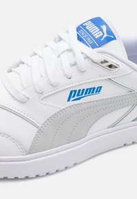 White leather sneaker with a grey accent, featuring a blue logo on the tongue and white laces. Textured surface with a smooth finish.