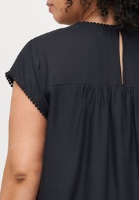 Blusa negra de manga corta, adornada con un dobladillo en forma de concha. Incluye una abertura en forma de llave en la parte posterior y tejido recogido para un ajuste relajado.