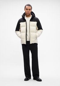 Letvægts, polstret jakke med en sort topsektion og cremet bund, med hætte og lynlås foran. Båret over en cremet sweater og sorte bukser.