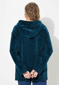 Tee-farbener Kapuzenpullover mit einem strukturierten, plüschigen Stoff, mit gerippten Bündchen und Saum, von hinten betrachtet. Die Hände sind hinter dem Rücken verschränkt.