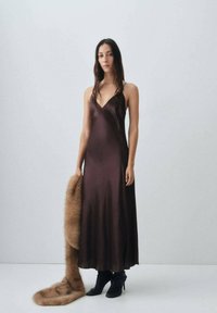 Braunes Satin-Cocktailkleid mit V-Ausschnitt und dünnen Trägern. Das Modell hält einen langen, hellbraunen Pelzschal. Schwarze, knöchellange, hohe Stiefel runden den Look ab.