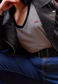 Osoba v černé kožené bundě, pruhované košili Levi’s, modrých džínách a širokém černém opasku s obdélníkovou sponou.