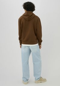 Personne aux cheveux bouclés portant un sweat à capuche marron, un jean bleu clair et des chaussures beiges, debout face à un fond uni gris clair.