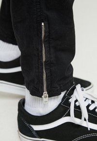 Svarta byxor med silverdragkedja vid vristerna över vita strumpor, kombinerat med svart- och vitrandiga snörskor.