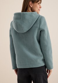 Veste en polaire bleu clair avec une capuche, présentant une surface texturée et une coupe décontractée, associée à un pantalon sombre.