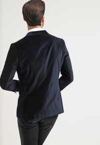 Blazer di velluto blu navy con vestibilità su misura, dotato di uno spacco singolo sul retro e tessuto liscio. Indossato sopra a jeans scuri.