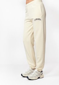 Kremowe dresy z elastycznym pasem, bocznymi kieszeniami i haftem "LAMANIA" w ciemnym kolorze na lewej udzie.