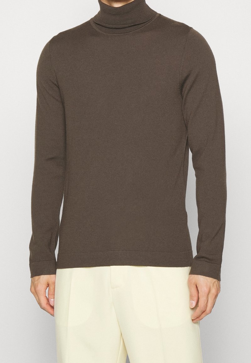Maglione a collo alto marrone realizzato in morbido tessuto a maglia, design aderente, maniche lunghe, colletto a coste, indossato sopra pantaloni di colore chiaro.
