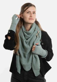 Jonge vrouw in een zwarte jas met lichtgroene gebreide handschoenen zonder vingers en een bijpassende gestructureerde sjaal, terwijl ze haar haar aanpast.