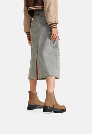 Stivali ankle in suede marrone con suola nera spessa, indossati con una gonna in denim azzurro chiaro con uno spacco sul retro e tasche laterali.