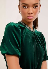 Zielono-szmaragdowy aksamitny top z bufkowymi rękawami, z plisowanym wzornictwem i ozdobnym dekoltem. Gładka tekstura i bogata głębia koloru.