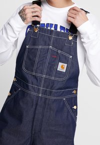 Denimoverall i mörkblått med kontrasterande vit sömnad, sidoknappar och en framlapp med en Carhartt-logotyp.