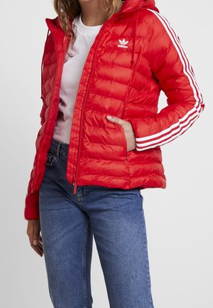 Persoon draagt een rood gewatteerd Adidas-jas met witte strepen op de mouwen, blauwe spijkerbroek en een wit shirt, hand in de jaszak.