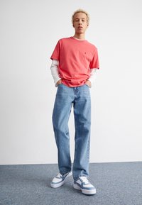 Joven con cabello rubio rizado que está de pie con las manos en los bolsillos, vistiendo una camiseta de manga corta color coral sobre una camisa blanca de manga larga, jeans azules y zapatillas blancas.