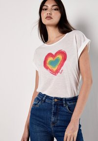 T-shirt blanc à manches courtes avec un design de cœur coloré incluant du rouge, du jaune, du vert et du bleu. Associé à un jean bleu taille haute.