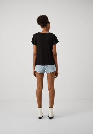 Top negro de manga corta, combinado con shorts de mezclilla deshilachados. La modelo lleva botas blancas hasta el tobillo. El fondo liso enfatiza el conjunto.