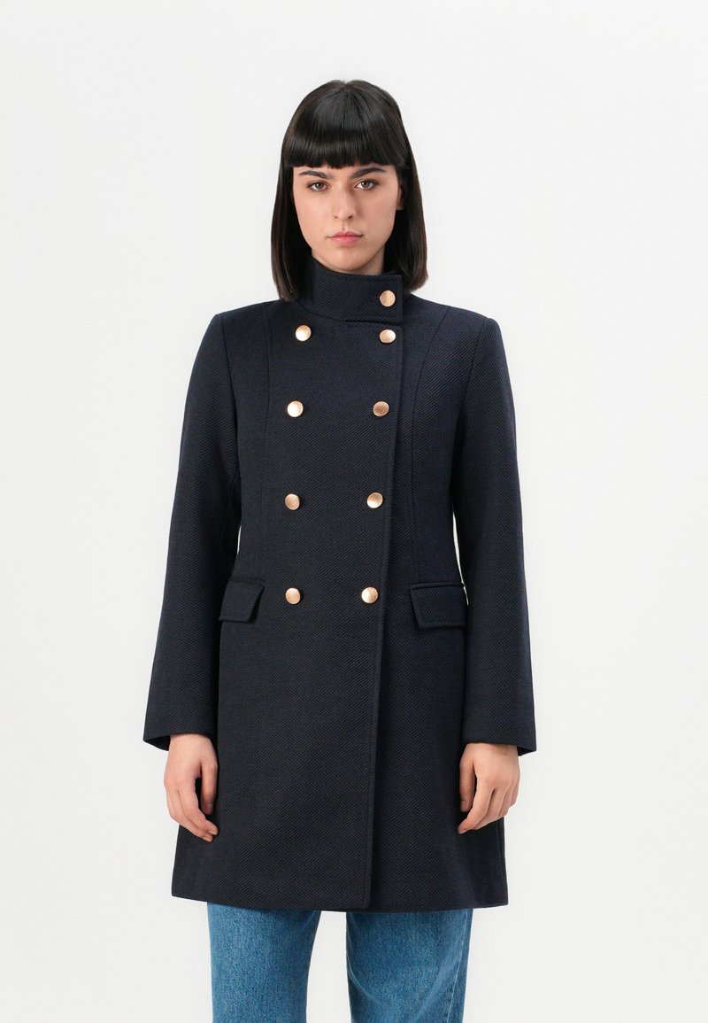 Femme portant un manteau noir ajusté à double boutonnage avec des boutons dorés, se tenant droite devant un fond blanc uni.