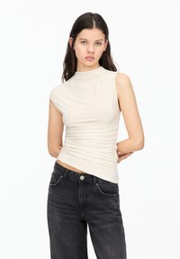 Top beige a spalla unica con un design aderente e una texture arricciata, abbinato a jeans in denim scuri a vita alta con tasche anteriori.