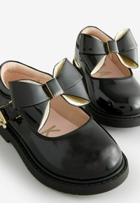 Scarpe in vernice nera con punta arrotondata, dotate di un fiocco decorativo e accenti in metallo dorato. L'interno è di un rosa chiaro con il brand.