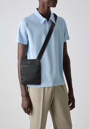 Schwarze Nylontasche mit Quergurt und Reißverschluss, mit Logodetail, getragen über einem hellblauen Poloshirt und beigen Hosen.
