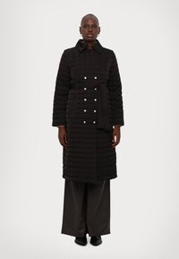 Melnā quiltota garā jaka ar jostiņu viduklī, sudraba pogu detaļām un apkakles izgriezumu. Iezīmē gludu tekstūru un taisnu piegriezumu.